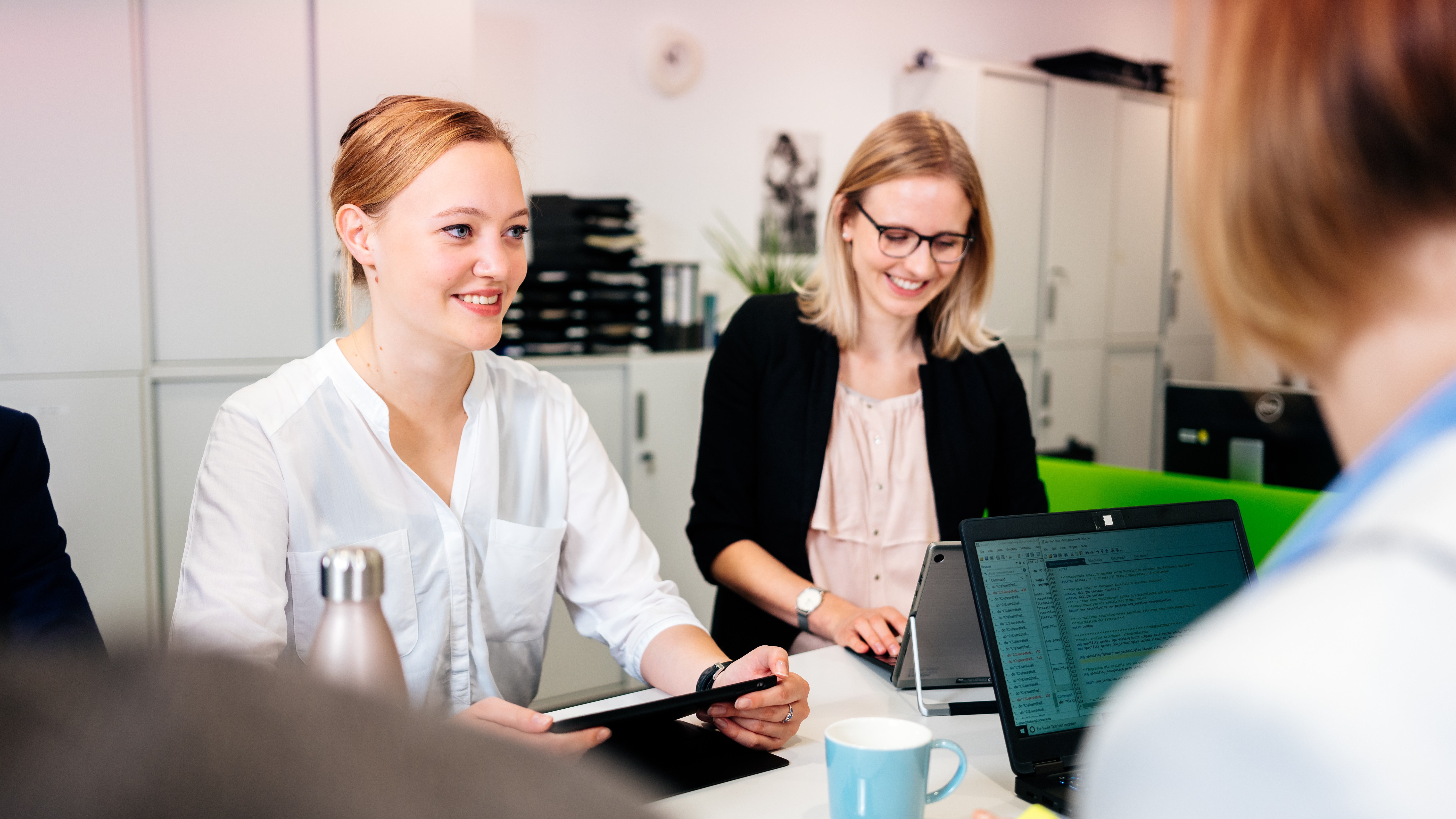 Drei Frauen sitzen an einem Arbeitsplatz mit Laptops und Tablet. 